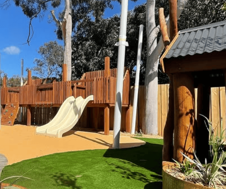 Wooden outdoor play structure with slide, kids’ playground equipment, and natural landscape at DG Play, outdoor recreational facility, family-friendly play area, adventure playground, safety surfacing, nature-inspired play zone.