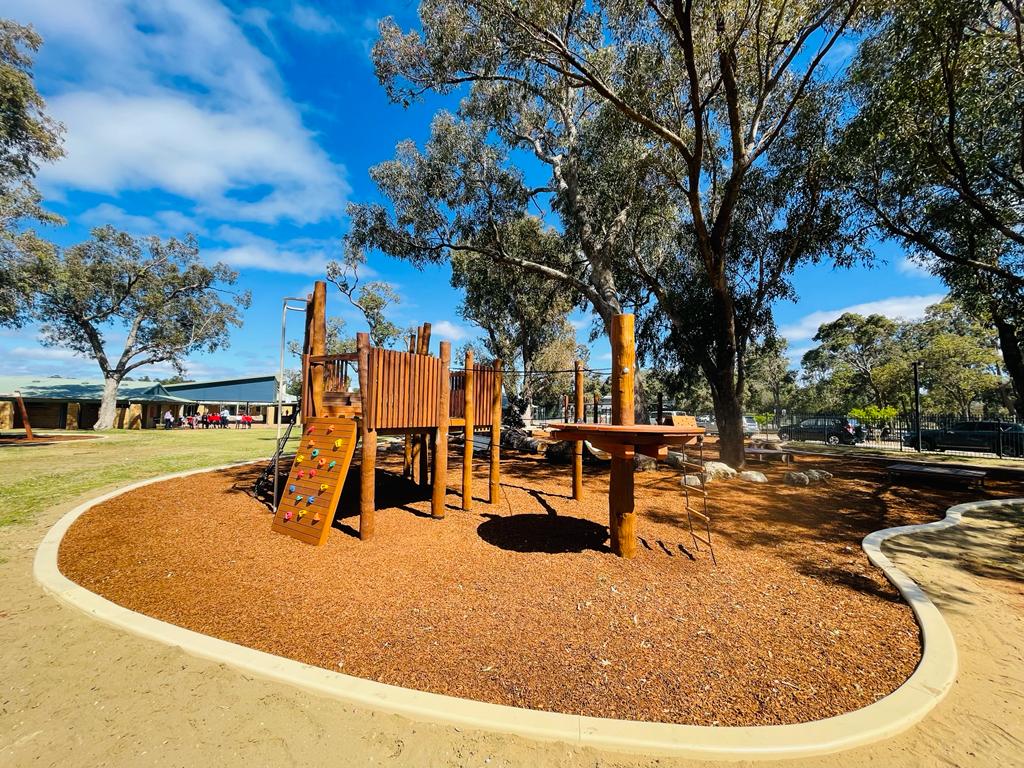Natural outdoor playground with wooden play structures and climbing wall, surrounded by tall trees and a sunny sky, providing a safe and engaging space for children.
