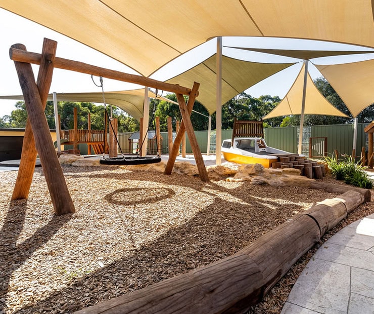 Natural outdoor playground with swings, sandbox, wooden climbing structures, and shade sails for children’s play and outdoor recreation.