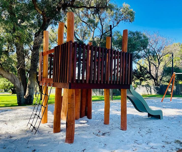 Climbing playground structure with slide and sand surface in a lush outdoor park setting.