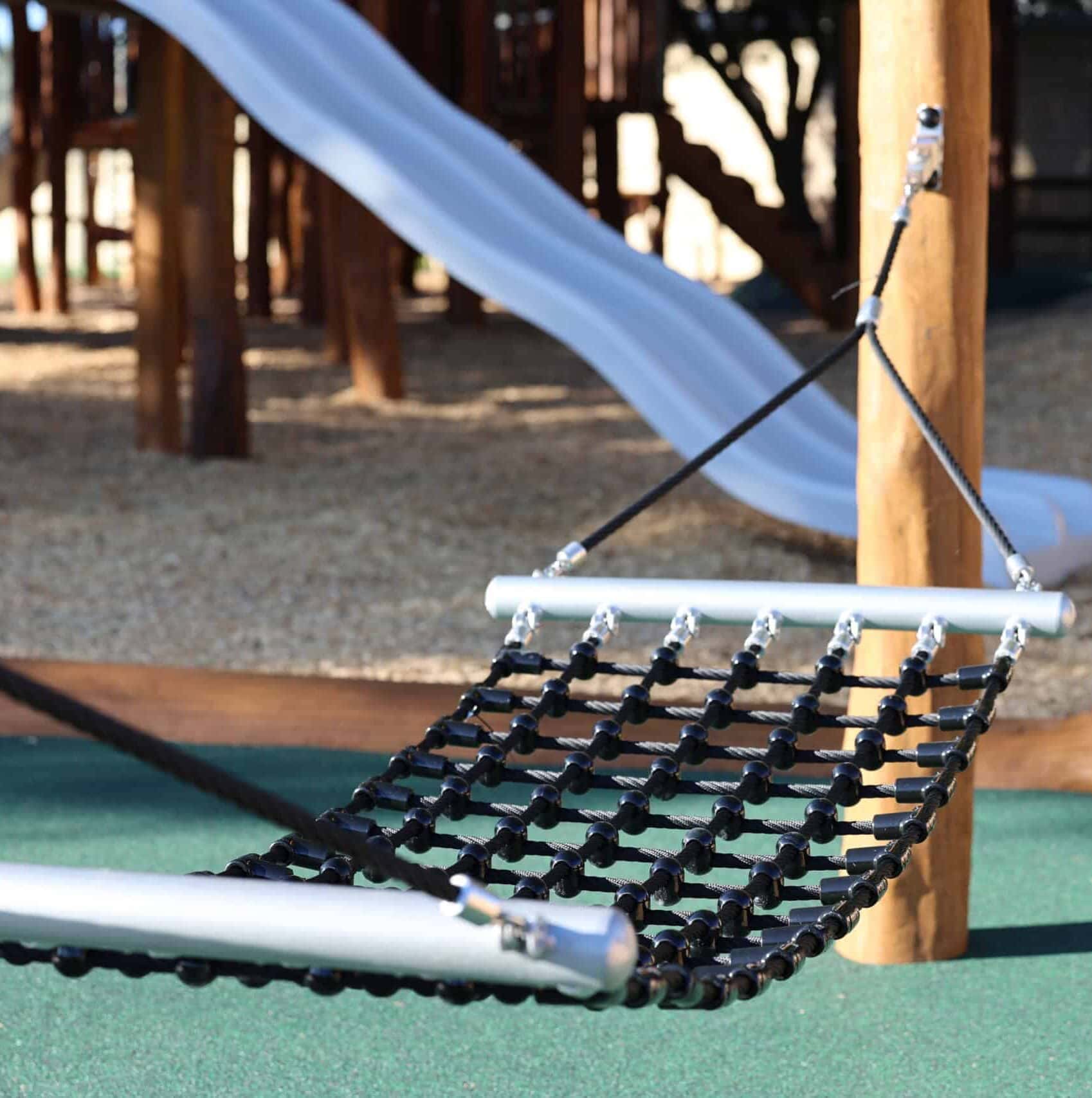 wilderness walk gallery Modern playground swing with a black net seat and metal frame, featuring a blue slide in the background for kids' outdoor fun and recreational activities.