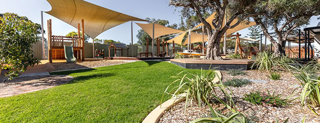 Elevated Platforms Home Bright outdoor playground with shaded areas, lush green lawn, and natural landscaping, perfect for children to play and enjoy in a safe, family-friendly environment.