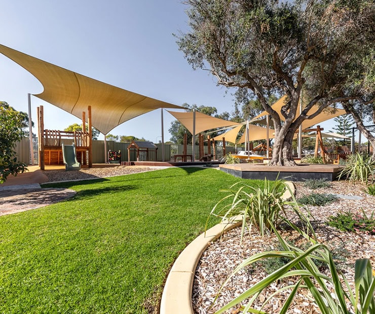 Bright outdoor backyard with shade sails, lush green lawn, mature trees, play structure, and landscaped garden for kids' play and relaxation.