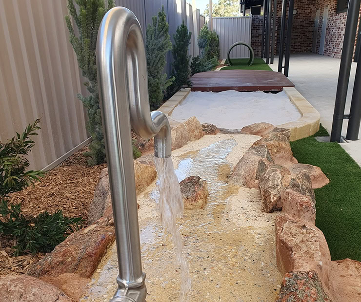 Natural rock water play feature with stainless steel spout and sandy stream, perfect for outdoor kids' activity area at DG Play outdoors.