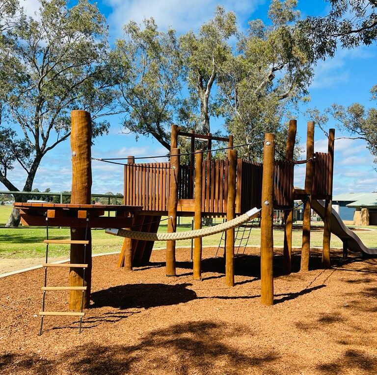 Bright and colorful wooden playground structure for children, featuring climbing areas, slides, and bridges, set in a sunny outdoor park with trees and blue sky.