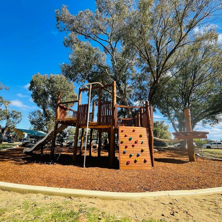 A vibrant outdoor playground featuring a wooden fort structure with slides, climbing walls, and swings, set amidst lush trees and a clear blue sky.