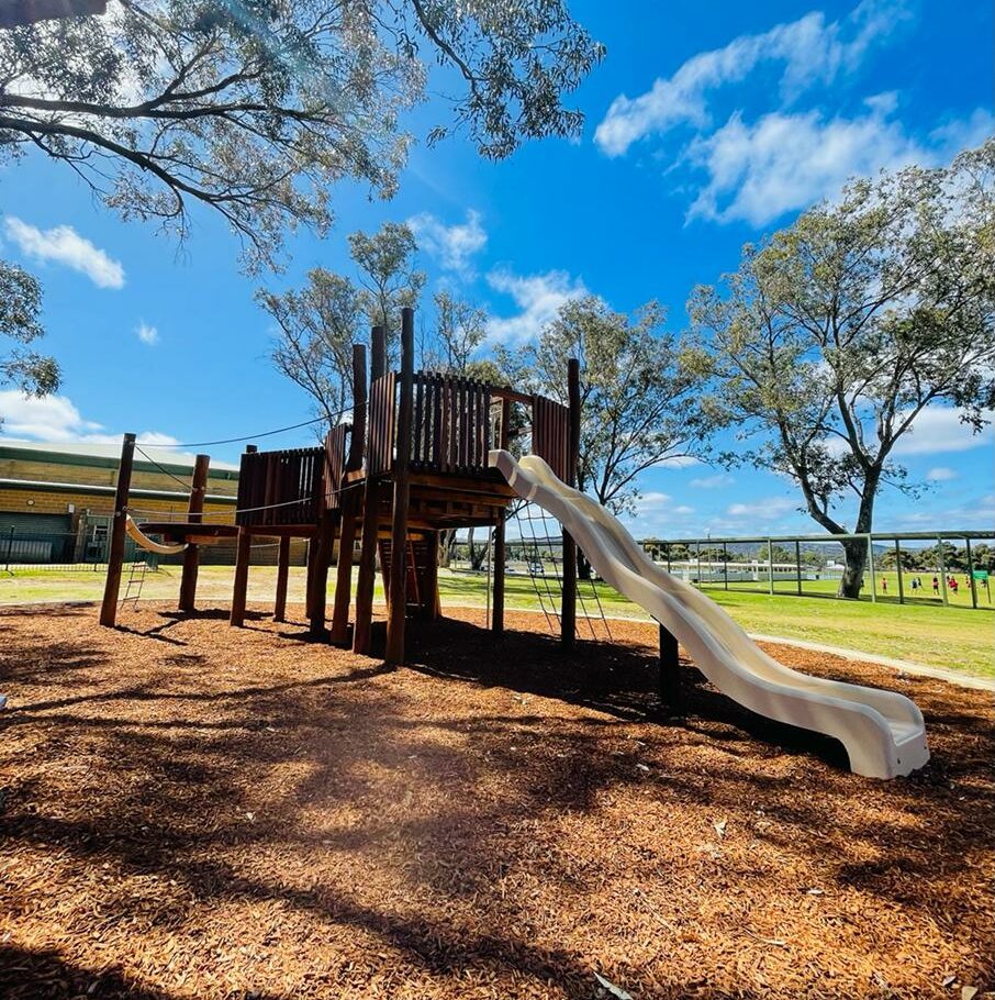 Wooden playground structure with slide at Big John Fort Park, featuring climbing areas and open space for children’s outdoor play.