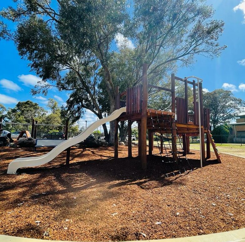 Playground with slide and climbing structure at Big John Fort, featuring a large tree providing shade, ideal for children's outdoor activities and family fun.
