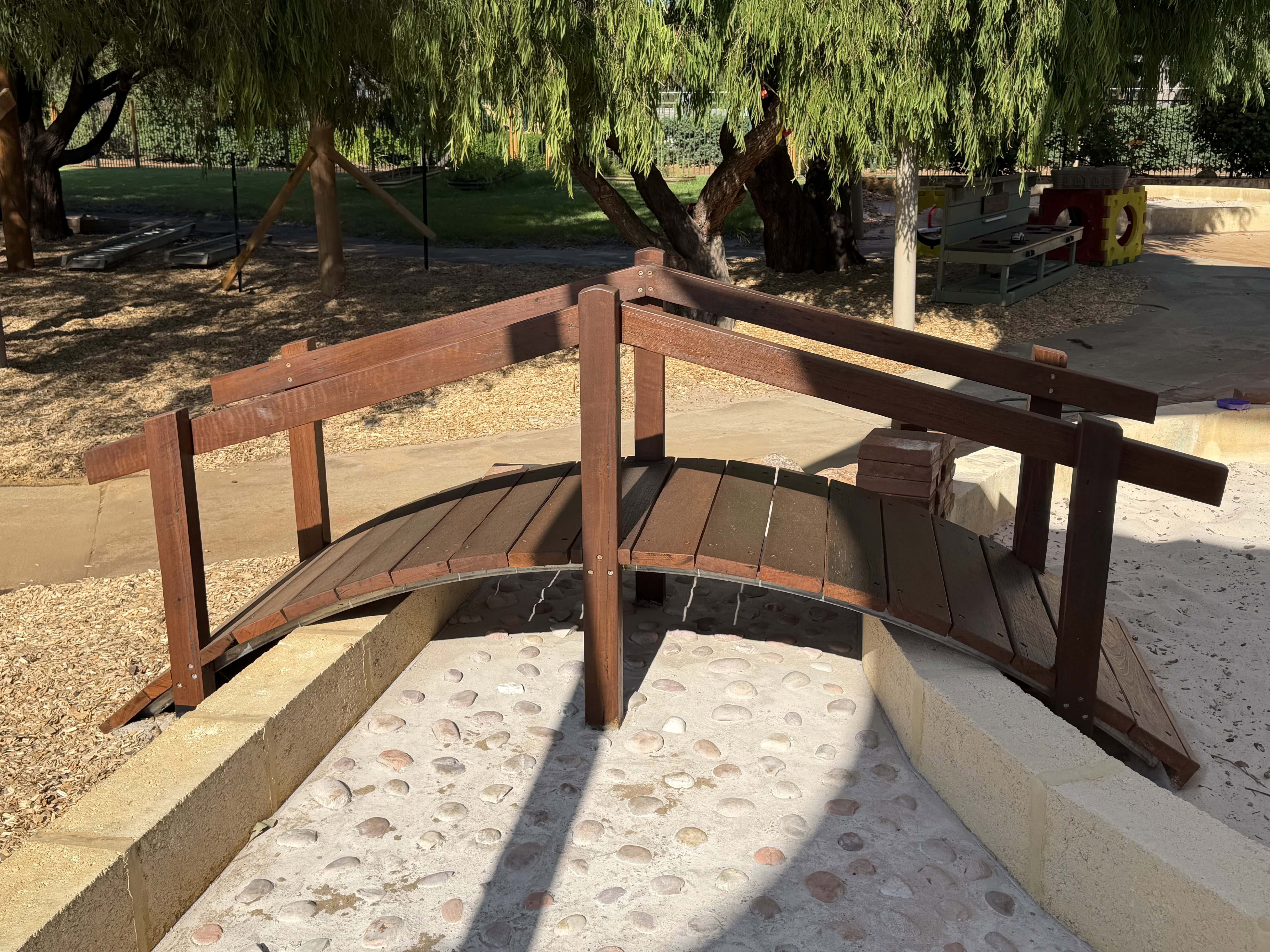 Wooden playground bridge with handrails in a park setting, surrounded by trees and sand.