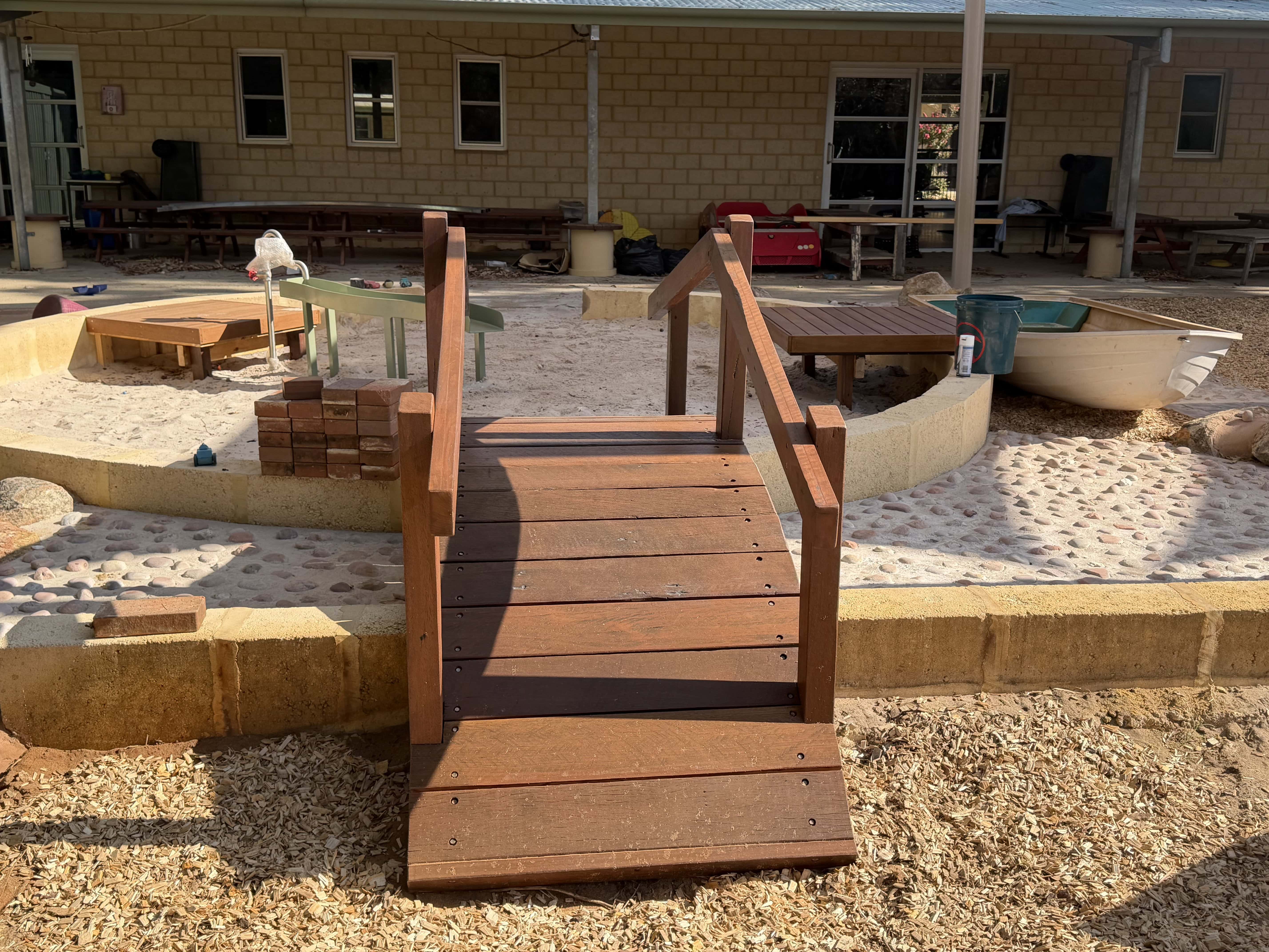 Colorful children's playground featuring a small wooden bridge, sandbox, and play area with boats and toys, set against a building with windows and outdoor seating.