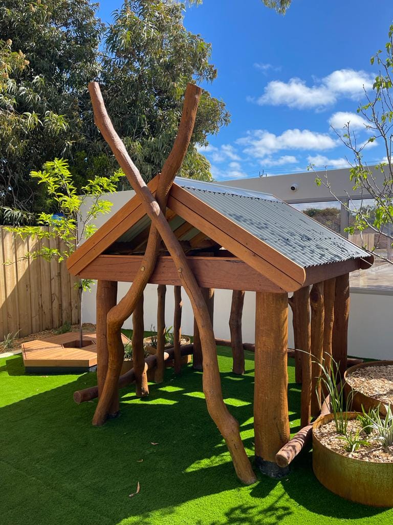 Wooden playhouse with natural logs and a metal roof, set in a backyard with green artificial grass and trees, under a blue sky.