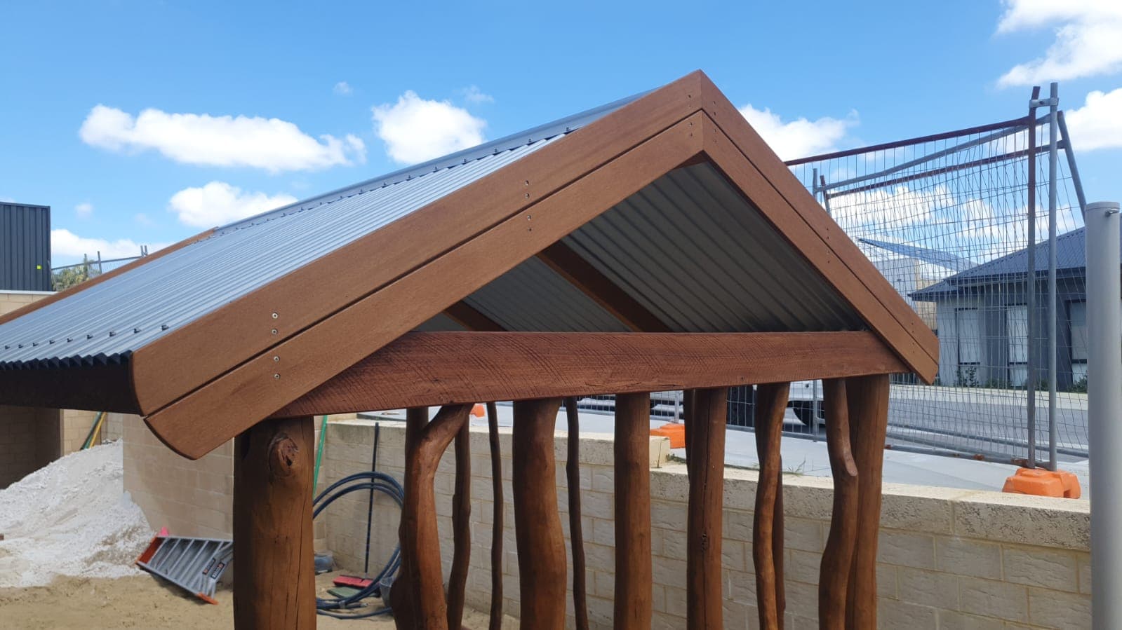 Small wooden playhouse with a sloped metal roof, supported by natural wood logs, set outdoors with a blue sky and clouds in the background.